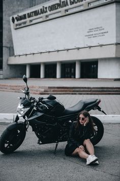 a woman sitting on the ground next to a motorcycle in front of a large building