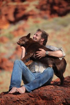 a man sitting on top of a red rock holding a brown dog in his arms