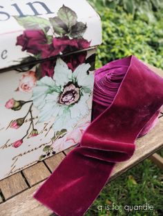 a purple ribbon is tied to a box on a wooden bench in front of some bushes