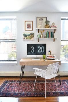 a desk with a computer on top of it in front of two windows and a rug