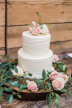 a white wedding cake with pink roses and greenery sits on a wooden table in front of wood planks