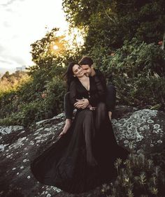 a man and woman sitting on top of a rock in the grass with their arms around each other