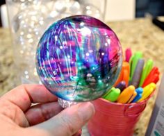 a hand holding a colorful glass ball in front of a cup filled with pens and markers