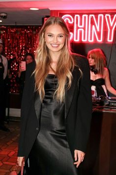 a woman standing in front of a neon sign at a party wearing a black dress and blazer