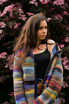 a woman wearing a multicolored crocheted cardigan standing in front of pink flowers