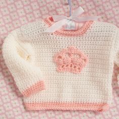 a pink and white knitted sweater with a crown on the front hanging from a hook