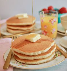 a stack of pancakes with butter and syrup being poured onto them on a table set for breakfast