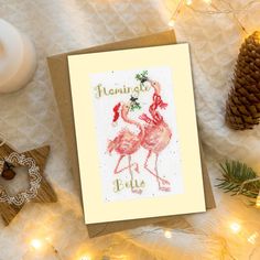 a cross stitch card with two flamingos on it next to pine cones and christmas lights