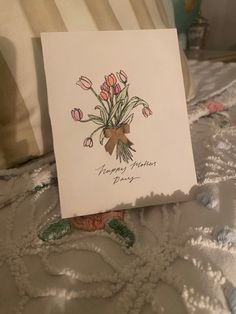 there is a card with flowers on it sitting on the edge of a bed next to a pillow