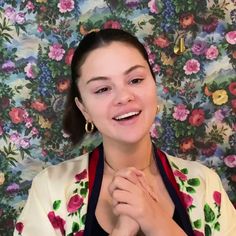 a woman in a floral shirt is smiling and holding her hands together while standing against a wall with flowers on it