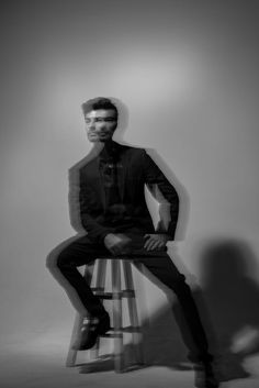 a man sitting on top of a wooden stool in front of a white wall with his legs crossed