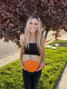 a pregnant woman holding an orange pumpkin in her belly while standing next to a tree