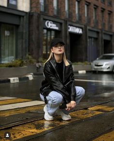 a woman sitting on the ground in front of a building wearing a black jacket and white sneakers