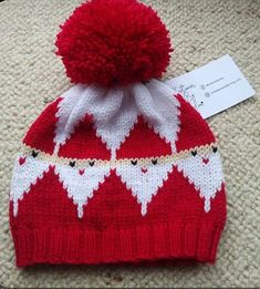 a red and white knitted beanie hat with hearts on it, next to a tag