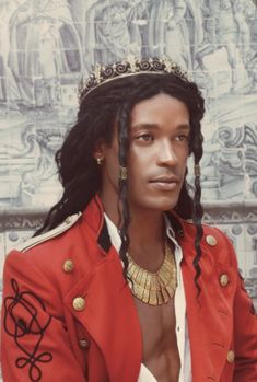 a man with dreadlocks wearing a red jacket and gold necklace, standing in front of a wall