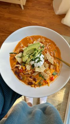 a person holding a bowl of soup with tortilla chips and avocado