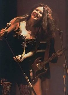 a woman with long hair holding a guitar and singing into a microphone while standing on stage