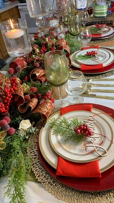 the table is set for christmas dinner with red and white plates, silverware, greenery and candlesticks