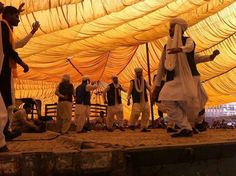 men in traditional garb dancing under a large yellow tent with people standing around it