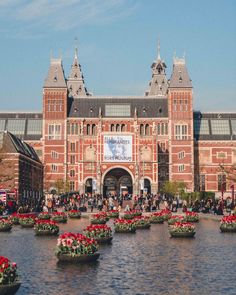 people are walking around in front of a large building with many flower pots on the water