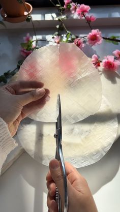 someone is cutting paper with scissors on a white table next to pink flowers and potted plants