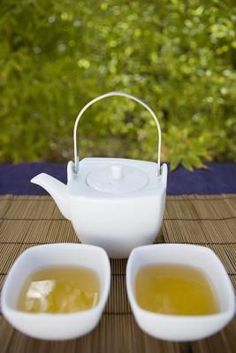 two white bowls filled with green tea sit on a bamboo mat next to a teapot