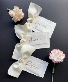 three ribbons with name tags on them sitting next to two pink carnations and one white flower