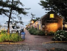 a street lined with parked cars next to trees and flowers at dusk, in front of a small wooden building