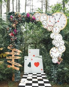 a checkerboard table topped with lots of clocks next to trees and flowers in the background