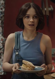 a woman in overalls holding a plate with food on it and looking at the camera