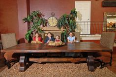 three children sitting at a table in front of a clock and potted plants on the floor