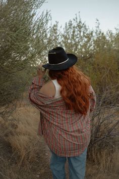 a woman with red hair wearing a plaid shirt and cowboy hat standing in the brush