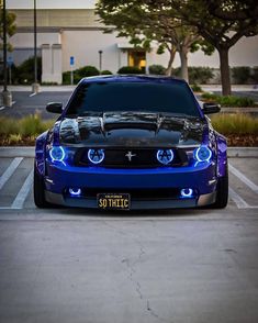 the front end of a blue mustang in a parking lot with its hood up and lights on