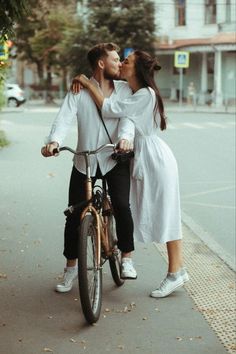 a man and woman kissing while riding a bike down the street with their arms around each other