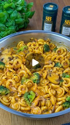 a pan filled with pasta and broccoli on top of a wooden table next to two cans of beer