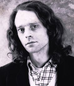 a black and white photo of a man with long hair wearing a plaid shirt, jacket and tie