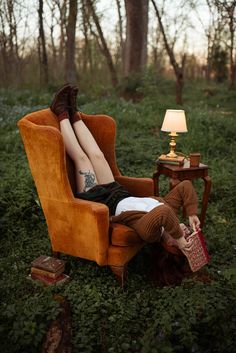 A woman sits in an orange velvet chair in the middle of the woods. Next to her is a side table with a glowing lamp and a coffee mug. She is surrounded by books, and is at peace amidst the forest as the sun sets into darkness. Best Props For Photoshoots, Photoshoot In Chair, Inside Outside Photography, Photoshoot In Home Ideas, Photograph Aesthetic Vintage, Fall Time Photoshoot, Painted Mines Photoshoot, Wood Photoshoot Ideas, Photography With Backdrops