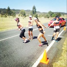 four people are running on the road in front of a car and orange traffic cones