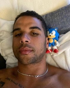 a shirtless man laying on top of a bed next to a sonic the hedge toy