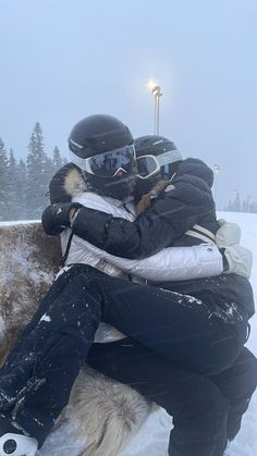 two people sitting on top of a snow covered ground with their arms around each other