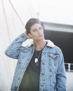 a young man wearing a denim jacket leaning against a wall with his hand on his head