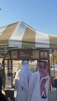 two men standing under a tent in front of other people and one man is wearing a headscarf