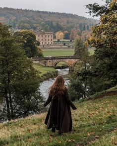 a woman walking down a hill next to a river with a bridge in the background