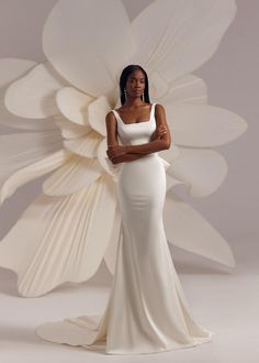 a woman in a white dress standing next to a large paper flower with her arms crossed