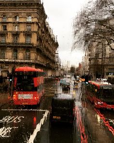 two red double decker buses driving down a street in the rain next to tall buildings