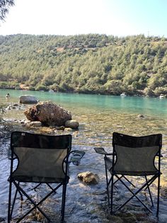 two chairs are sitting in the water near some rocks and trees, one is empty