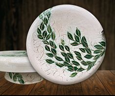 two white plates with green leaves painted on them sitting on a wooden table next to each other