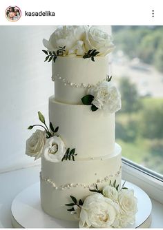 a wedding cake with white flowers and pearls on the bottom tier is sitting in front of a window