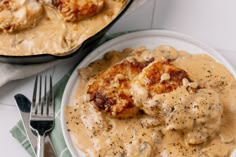 a white plate topped with chicken covered in gravy next to a skillet