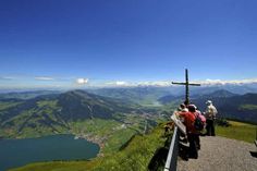 people standing at the top of a hill looking down on a lake and mountains in the distance